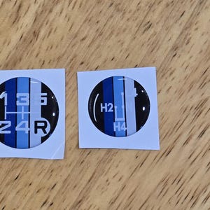 May include: Two white stickers with blue and black circular decals. One decal shows a gear shift pattern with the numbers 1, 2, 3, 4, and R. The other decal shows a gear shift pattern with the letters H2 and H4.