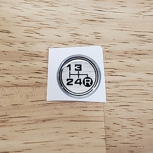 May include: A silver and black gear shift pattern sticker with the numbers 1, 2, 3, 4, and the letter R.