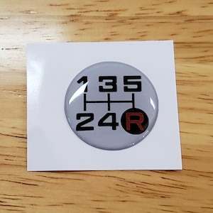 Puede incluir: Una pegatina circular gris con un patrón de cambio de marchas negro con los números 1, 2, 3, 4 y 5, y la letra "R" en rojo. La pegatina está sobre un cuadrado blanco.