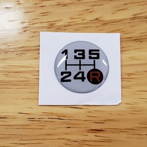 May include: A silver-colored, round, 5-speed gear shift pattern sticker with black numbers and a red 'R' for reverse.