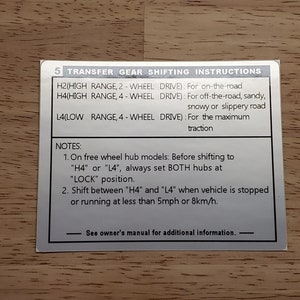 May include: A silver metal instruction label with black text explaining how to use the transfer gear on a vehicle. The label includes instructions for using the H2, H4, and L4 ranges, as well as notes about free wheel hub models. The label also includes the text "See owner's manual for additional information."