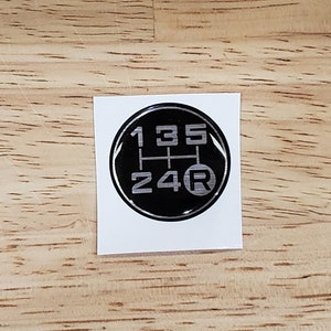 May include: A black and silver 5-speed manual transmission shift pattern sticker. The numbers 1, 3, and 5 are on the top row, 2 and 4 are on the bottom row, and "R" is to the right of the 4.