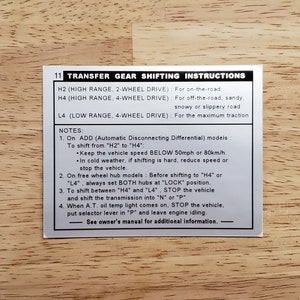 May include: A silver sticker with black text provides instructions for shifting a transfer gear. The sticker includes information about the different drive modes: H2 (High Range, 2-Wheel Drive), H4 (High Range, 4-Wheel Drive), and L4 (Low Range, 4-Wheel Drive). The sticker also includes notes about shifting between the different drive modes.