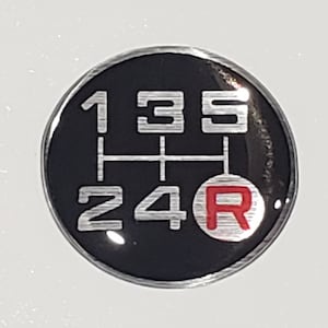 May include: A black round sticker with a silver gear shift pattern. The numbers 1, 3, 5, 2, 4 are in silver with a red circle with the letter R in the center.