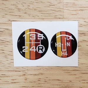 May include: Two round, black, glossy stickers with white numbers and letters on them. The stickers are designed to look like a car's gear shift pattern. One sticker shows the numbers 1, 3, 5, 2, 4, and R. The other sticker shows the letters H2, N, and H4.