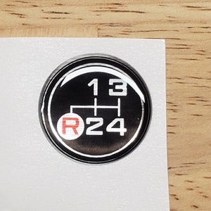 May include: A black and silver round sticker with a white gear shift pattern and the numbers 1, 2, 3, and 4. The letter "R" is in red.