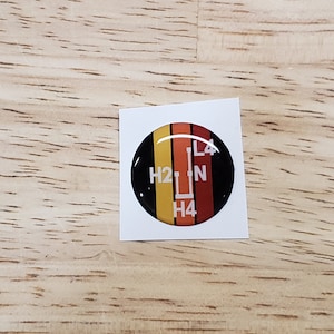 May include: A round, black, white, yellow, and red sticker with a gear shift pattern. The sticker has the letters H2, N, L4, and H4 printed on it.