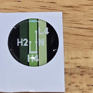 May include: A round, black and green sticker with the text "H2", "L4", "N", and "H4". The sticker is on a white, rectangular piece of paper. The background is a wooden surface.