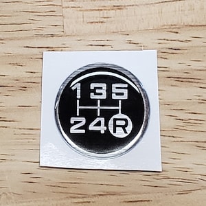 May include: A black and silver round sticker with a 5-speed manual transmission gear shift pattern. The numbers 1, 3, and 5 are at the top of the pattern, 2 and 4 are at the bottom, and R is to the right of the pattern.