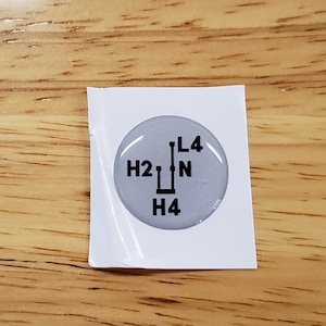 May include: A round, silver-colored sticker with black text and a gear shift pattern. The text reads "H2", "N", "L4", and "H4".