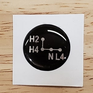 May include: A black, circular sticker with a glossy finish. The sticker features white text and a diagram indicating gear positions: H2, H4, N, and L4. The sticker is on a white, square backing.