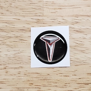 May include: A black and silver Tesla logo sticker with a red outline. The sticker is round and has a glossy finish.