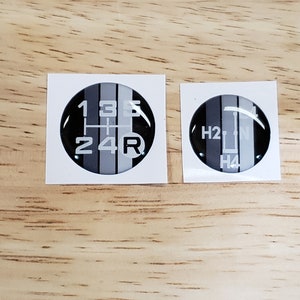 May include: Two black and white round stickers with gear shift patterns. The first sticker shows the numbers 1, 3, 5, 2, 4, and R. The second sticker shows the letters H, 2, N, and H4.