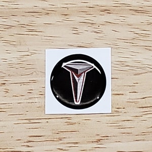 May include: A black and white Tesla logo sticker with a red outline. The sticker is round and has a glossy finish.