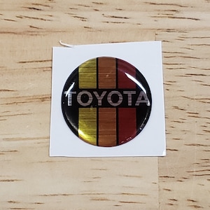 May include: A round, black and silver Toyota logo sticker with gold and red stripes. The sticker is on a white background.