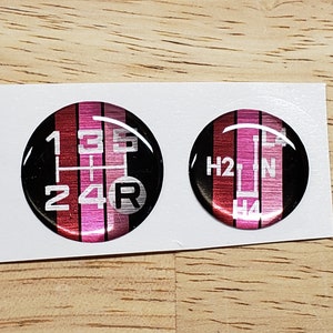 May include: Two round, black and pink car gear shift pattern stickers. One sticker shows the pattern for a 5-speed manual transmission with reverse. The other sticker shows the pattern for a 4-speed automatic transmission with a neutral position.