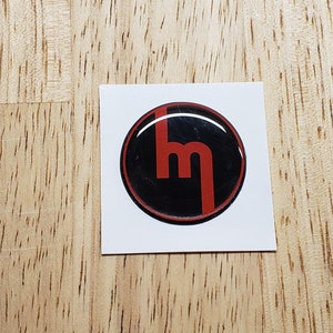 May include: A red and black round emblem with the letter "M" in the center. The emblem is glossy and has a raised design.