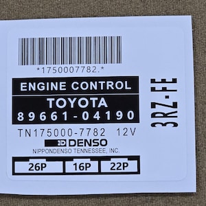 May include: A white sticker with black text and a barcode. The sticker reads "ENGINE CONTROL TOYOTA 89661-04190 TN175000-7782 12V DENSO NIPPONDENSO TENNESSEE, INC." with additional codes and "3RZ-FE".