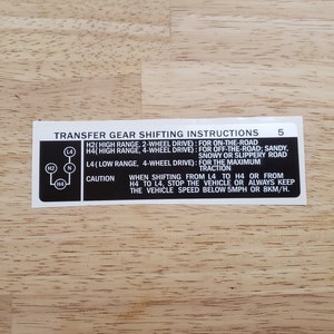 May include: A rectangular sticker with black text on a white background, providing transfer gear shifting instructions. The sticker includes diagrams and text detailing gear positions and driving conditions. The sticker is on a wooden surface.