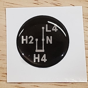 May include: Black round sticker with white text and a diagram showing the gear shift positions for a vehicle. The text reads "H2", "N", "H4", and "L4".