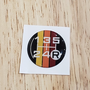 May include: A black and white 5-speed manual transmission gear shift pattern sticker with a red, orange, and yellow stripe design. The sticker has the numbers 1, 2, 3, 4, 5, and the letter R.