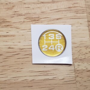 May include: A gold-colored round sticker with a white gear shift pattern and the numbers 1, 3, 5, 2, 4, and the letter R.