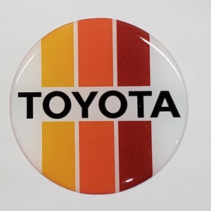 May include: A white round sticker with the word "TOYOTA" in black letters. The sticker has three vertical stripes in yellow, orange, and red.