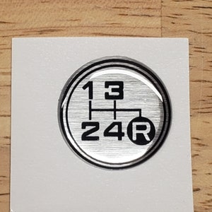 May include: A silver-colored round sticker with a black gear shift pattern. The numbers 1, 2, 3, 4, and the letter R are displayed on the sticker.