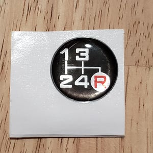 May include: A black and white gear shift pattern sticker with a red "R" for reverse. The sticker is on a white background.