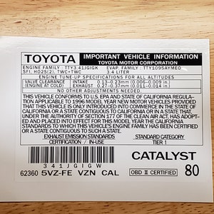 May include: A white sticker with black text detailing vehicle information for a 1996 Toyota. The sticker includes the engine family, emissions standards, and OBD II certification. The text "CATALYST" is printed in black on a white background.