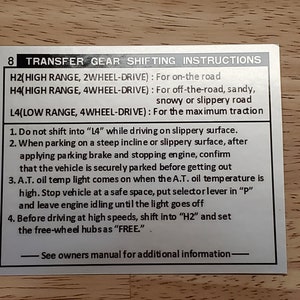 May include: A silver metal sign with black text provides instructions for a vehicle's transfer gear shifting. The sign includes the text "H2 (High Range, 2Wheel-Drive): For on-the-road", "H4 (High Range, 4Wheel-Drive): For off-the-road, sandy, snowy or slippery road", and "L4 (Low Range, 4Wheel-Drive): For the maximum traction". The sign also includes a list of four instructions for using the transfer gear.