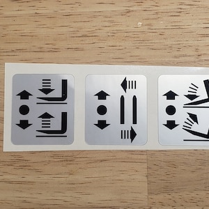May include: Three silver stickers with black arrows and symbols indicating different seat adjustments. The first sticker shows up and down arrows with a symbol of a seat moving up and down. The second sticker shows arrows pointing left and right with a symbol of a seat moving left and right. The third sticker shows arrows pointing up and down with a symbol of a seat reclining.