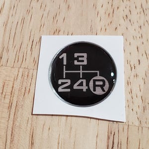 May include: A black, circular gear shift pattern sticker with white text. The sticker shows the gear positions: 1, 2, 3, 4, and R (reverse). The sticker is on a white backing, set against a wooden surface.