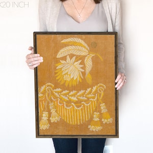 May include: A framed print of a vintage floral design in yellow and white on a mustard yellow background. The design features a stylized flower with leaves and a decorative border.