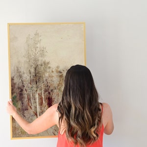 May include: A woman is hanging a framed painting on a white wall. The painting is a watercolor of a forest scene with trees and a light brown background.