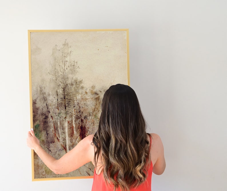 May include: A woman is hanging a framed painting on a white wall. The painting is a watercolor of a forest scene with trees and a light brown background.