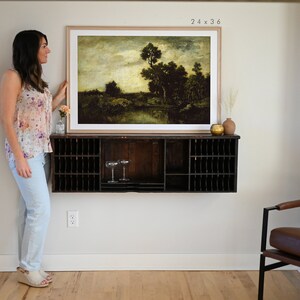 May include: A framed landscape painting of a forest scene with a river in the foreground. The painting is in a light wood frame and is hanging on a white wall above a dark wood shelf. The painting is 24 inches by 36 inches.
