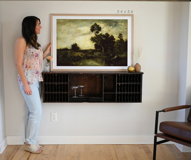 May include: A framed landscape painting of a forest scene with a river in the foreground. The painting is in a light wood frame and is hanging on a white wall above a dark wood shelf. The painting is 24 inches by 36 inches.