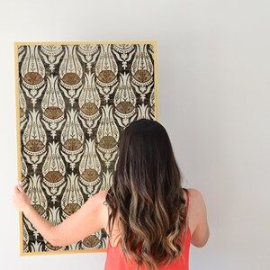 May include: A woman is hanging a framed print with a black and white floral pattern. The print has a gold frame and features a repeating pattern of stylized flowers.