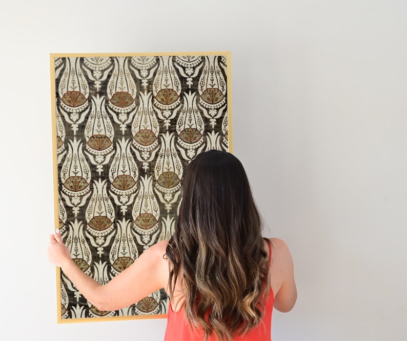 May include: A woman is hanging a framed print with a black and white floral pattern. The print has a gold frame and features a repeating pattern of stylized flowers.