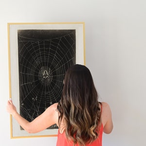 May include: A framed black and white print of a spider web with a spider in the center. The print is hanging on a white wall. A person is adjusting the frame.