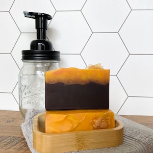May include: Two bars of handmade soap in a bamboo tray, with a mason jar soap dispenser. The top soap bar is dark brown with an orange top, and the bottom bar is orange with a decorative element. A gray towel is underneath the tray.