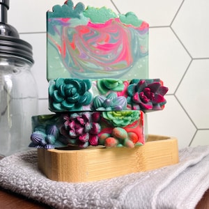 May include: Three bars of handmade soap with a succulent theme. The top bar has a green top with pink and purple swirls. The middle and bottom bars are decorated with colorful succulent designs. The soaps are displayed on a wooden soap dish.
