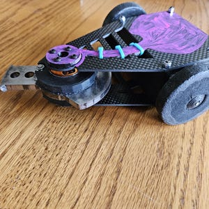 May include: A black and purple robot with a carbon fibre body and a purple and black motor. The robot has two black wheels and a purple and black spinning weapon.