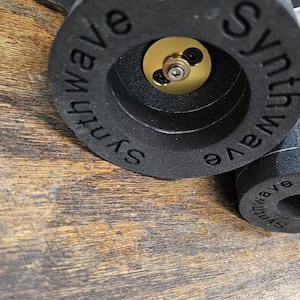 Puede incluir: Primer plano de un objeto redondo negro con el texto "Synthwave" escrito en él en un patrón circular. El objeto tiene un centro dorado con un pequeño tornillo.