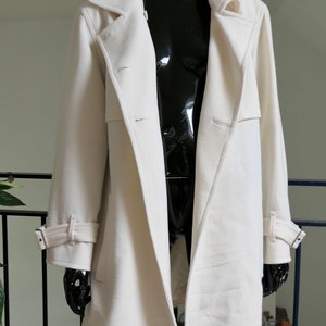 May include: A long, off-white coat with a classic design. The coat features a notched collar, button closures, and adjustable cuffs. The coat is displayed on a black mannequin, showcasing its tailored silhouette and details. A stylish outerwear piece.