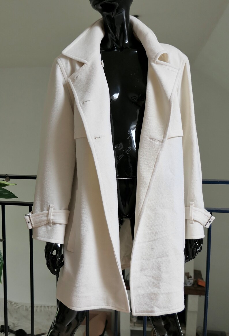 May include: A long, off-white coat with a classic design. The coat features a notched collar, button closures, and adjustable cuffs. The coat is displayed on a black mannequin, showcasing its tailored silhouette and details. A stylish outerwear piece.