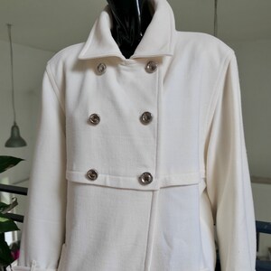May include: Cream-colored double-breasted coat with silver-toned buttons. The coat features a wide collar, a tailored fit, and a belt detail on the sleeves. The coat is displayed on a black mannequin.