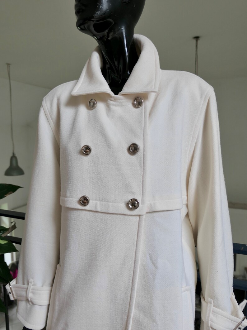 May include: Cream-colored double-breasted coat with silver-toned buttons. The coat features a wide collar, a tailored fit, and a belt detail on the sleeves. The coat is displayed on a black mannequin.