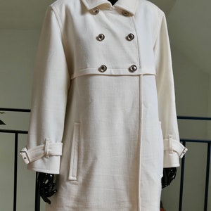 May include: Off-white double-breasted coat with silver-tone buttons. The coat features a notched collar, long sleeves with adjustable cuffs, and two front pockets. The coat is displayed on a black mannequin.